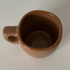 Pode incluir: Uma caneca de cer&acirc;mica com um esmalte castanho brilhante e salpicado. A caneca apresenta uma forma arredondada e org&acirc;nica com uma pega grossa e resistente fixada na lateral. O interior &eacute; liso e profundo, com o mesmo acabamento castanho.