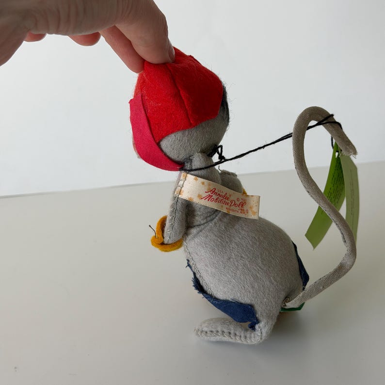 K&ouml;nnte beinhalten: Eine Vintage-Mauspuppe in Grau mit roter M&uuml;tze und langem, gebogenem Schwanz. Die Puppe hat einen gelben Gegenstand in der Hand und ein Etikett mit der Aufschrift "Annalee Mobilitee Doll". Die Maus hat einen blauen Flicken auf dem R&uuml;cken.