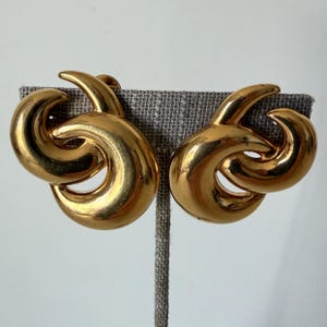 May include: A pair of gold-toned clip-on earrings. Each earring features a looped design with a polished finish. The earrings are displayed on a gray fabric surface.