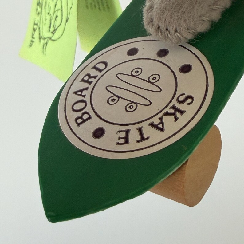 K&ouml;nnte beinhalten: Ein gr&uuml;nes Spielzeug-Skateboard mit Holzsockel und einem runden Aufkleber mit der Aufschrift "BOARD SKATE". Der Aufkleber zeigt zwei Skateboard-Illustrationen. Im Hintergrund ist ein hellgr&uuml;nes Etikett zu sehen.