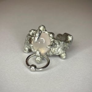 Könnte beinhalten: Ein silberfarbener Ring mit einem strukturierten, organischen Design, der einen hellrosa Edelstein aufweist. Ein separater kleiner Silberring mit Kugelverschluss ist ebenfalls vorhanden. Der Schmuck ist vor einem weißen Hintergrund platziert.