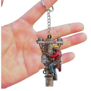 May include: A keychain featuring an electrician working on a utility pole. The charm has a detailed illustration of a lineman in a red jacket and yellow helmet. The text "YOUR NAME" is above the pole.