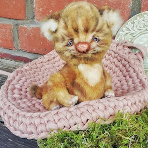 Op de afbeelding: Een kleine, pluche tijgerwelpfiguur in bruin en beige met blauwe ogen en een getextureerde neus, zittend in een ronde, lichtroze mand van grof gebreide stof. De welp heeft pluizige oren en een zachte vacht met donkerbruine strepen.