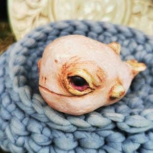 Peut inclure: Une sculpture fantaisiste, rose pâle, représentant une créature aux grands yeux expressifs et à la petite bouche. La sculpture repose dans un panier en tricot bleu clair et épais. Les yeux de la créature sont rouge foncé avec des reflets dorés.