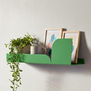 Może przedstawiać: Nowoczesna scena wnętrza z zieloną półką na ścianie z roślinami i oprawionymi dziełami sztuki. Drewniana ławka mieści książki i małą doniczkę z sukulentem. Pokój jest jasny, tworząc minimalistyczną estetykę.