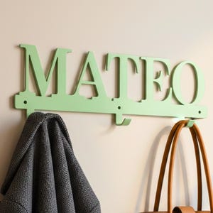 Op de afbeelding: Een mintgroene houten wandhaak met de naam "MATEO" in hoofdletters. Een grijze jas hangt aan de linkerhaak en een bruine leren draagtas aan de rechterhaak. De muur is neutraal beige.