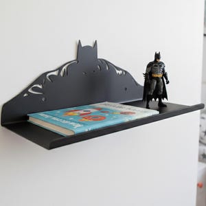 Op de afbeelding: Zwart metalen plank met een Batman silhouet. Een kinderboek met een blauwe kaft en een Batman-figuur staan op de plank. De plank is aan een witte muur bevestigd.