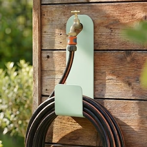 Może przedstawiać: Miętowy zielony uchwyt na wąż ogrodowy zamontowany na drewnianej ścianie. Uchwyt ma zakrzywioną konstrukcję z górną częścią na kran i dolną częścią do trzymania zwiniętego czarno-pomarańczowego węża. Kran jest mosiężny.