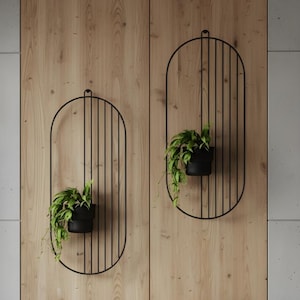 May include: Two black metal wall planters with oval frames and vertical bars, each holding a potted green plant. The planters are mounted on a wooden wall. A grey sofa with a geometric patterned cushion and a cream-coloured throw blanket is in the foreground.