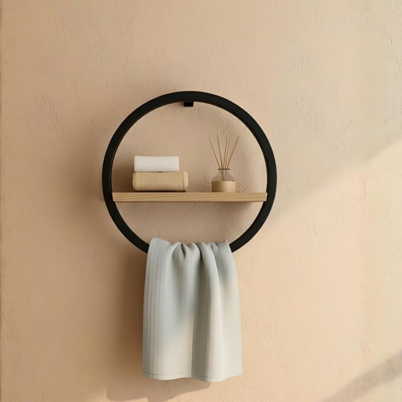 May include: A black circular wall shelf with a wooden shelf and a towel bar. The shelf holds a rolled white towel, a rolled beige towel, and a small wooden diffuser with reeds. A light gray towel hangs from the bottom.