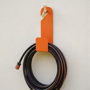 Puede incluir: Un soporte de manguera de metal naranja con un grifo de lat&oacute;n fijado a una pared blanca. Una manguera de jard&iacute;n negra y naranja est&aacute; enrollada y sujeta por el soporte. La manguera est&aacute; lista para usar en el jard&iacute;n.