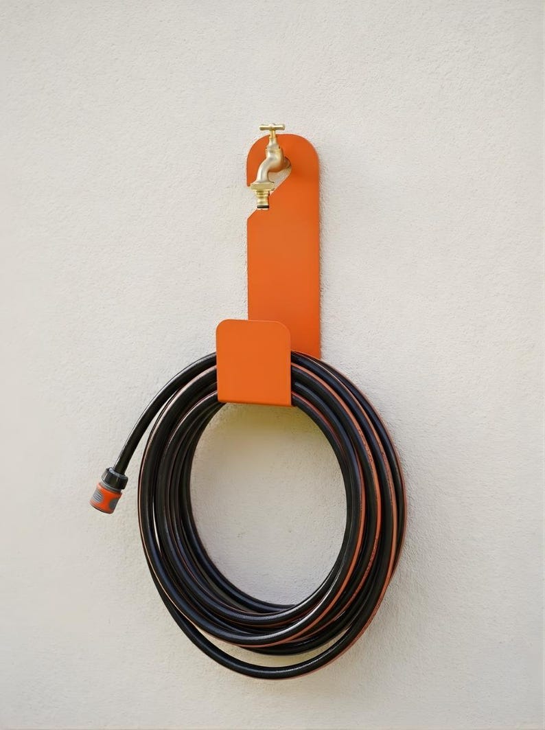Puede incluir: Un soporte de manguera de metal naranja con un grifo de lat&oacute;n fijado a una pared blanca. Una manguera de jard&iacute;n negra y naranja est&aacute; enrollada y sujeta por el soporte. La manguera est&aacute; lista para usar en el jard&iacute;n.