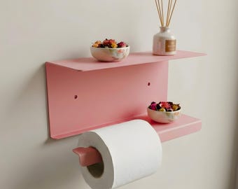 May include: A pink metal wall shelf with two levels, holding two small floral bowls, a white ceramic diffuser, and a roll of toilet paper with a pink holder. The shelf is mounted on a white wall.