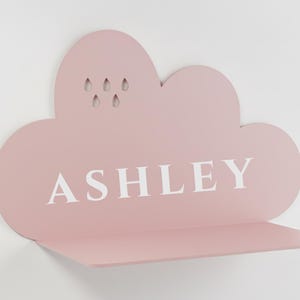 Op de afbeelding: Een roze, wolk-vormige plank met de naam "ASHLEY" in witte letters. De plank heeft een kleine plank en druppelvormige uitsparingen. Gemonteerd op een witte muur.