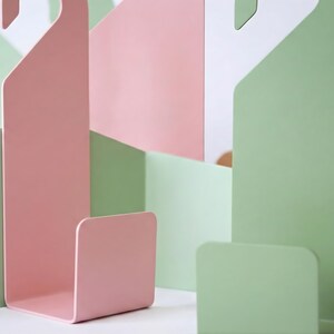 Puede incluir: Composici&oacute;n abstracta de formas met&aacute;licas en colores pastel. La imagen presenta una estructura rosa en forma de gancho y varias formas rectangulares verdes y rosas. El dise&ntilde;o es minimalista y moderno, centrado en el color y la forma.