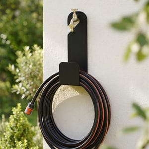 Puede incluir: Un soporte de manguera de metal negro montado en una pared blanca, con una manguera negra y naranja enrollada. Un grifo de lat&oacute;n est&aacute; fijado al soporte. El follaje verde es visible en el fondo.