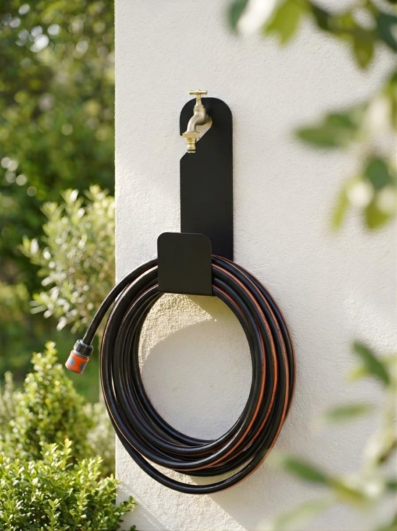 Puede incluir: Un soporte de manguera de metal negro montado en una pared blanca, con una manguera negra y naranja enrollada. Un grifo de lat&oacute;n est&aacute; fijado al soporte. El follaje verde es visible en el fondo.