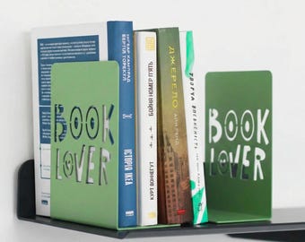 May include: Green metal bookends with the words "BOOK LOVER" cut out. Books of various colors and sizes are placed between the bookends. The books have titles in different languages.