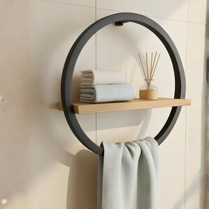 May include: A modern, circular wall-mounted towel rack with a black metal frame. It features a light wood shelf holding folded towels and a glass diffuser. A light green towel hangs from the bottom ring. The rack is mounted on a beige tiled wall.