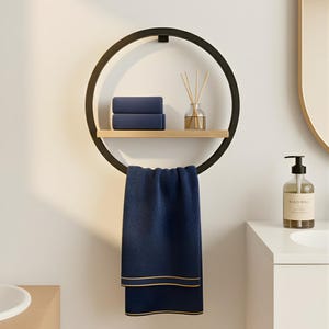 May include: A modern black metal towel rack with a circular design. It features a wooden shelf holding two folded navy blue towels and a glass diffuser. A navy blue towel with gold trim hangs below.