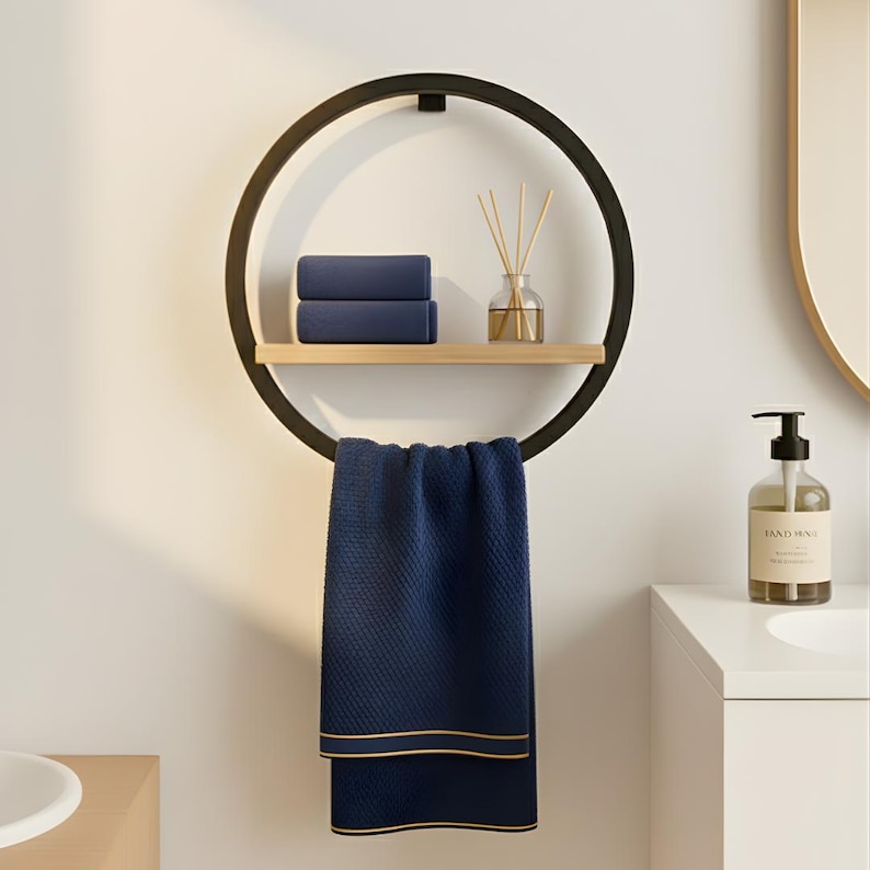 May include: A modern black metal towel rack with a circular design. It features a wooden shelf holding two folded navy blue towels and a glass diffuser. A navy blue towel with gold trim hangs below.