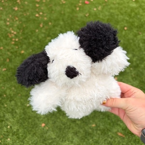 May include: A plush stuffed animal shaped like a dog with white fur and black patches on the ears and around the eyes. The toy features a soft, textured material, a small black nose, and two dark eyes, creating a simple, rounded appearance.