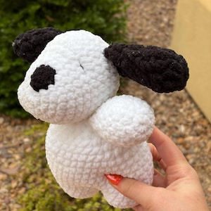 Puede incluir: Un juguete de perro de ganchillo hecho a mano en blanco con detalles en negro. El perro tiene orejas negras y una mancha negra sobre un ojo. El juguete está hecho de hilo suave y texturizado. El cuerpo del perro es redondo y afelpado.