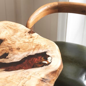 May include: Close-up of a live edge wooden table with a natural, organic shape. The wood grain is visible, with a mix of light and dark brown tones and a dark red resin-filled section. A curved wooden chair back is in the background.