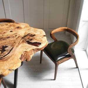 May include: A live edge wooden table with a natural, light brown finish and dark accents. A wooden chair with a curved back and a dark green leather seat is next to the table. The table has black metal legs.