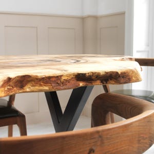 May include: A live edge wooden table with a natural, light brown finish. The table has a thick, irregular edge and is supported by a black metal V-shaped base. Wooden chairs surround the table.