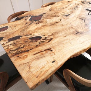 May include: A close-up of a wooden table with a natural edge. The tabletop features a light-colored wood grain with dark brown and reddish-brown accents. The table is surrounded by wooden chairs with black leather seats.