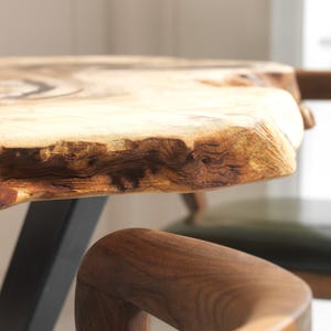 May include: A close-up of a wooden table with a natural edge. The table has a light brown surface with darker brown details. A wooden chair with a curved armrest is in the foreground. The table is supported by a black metal leg.