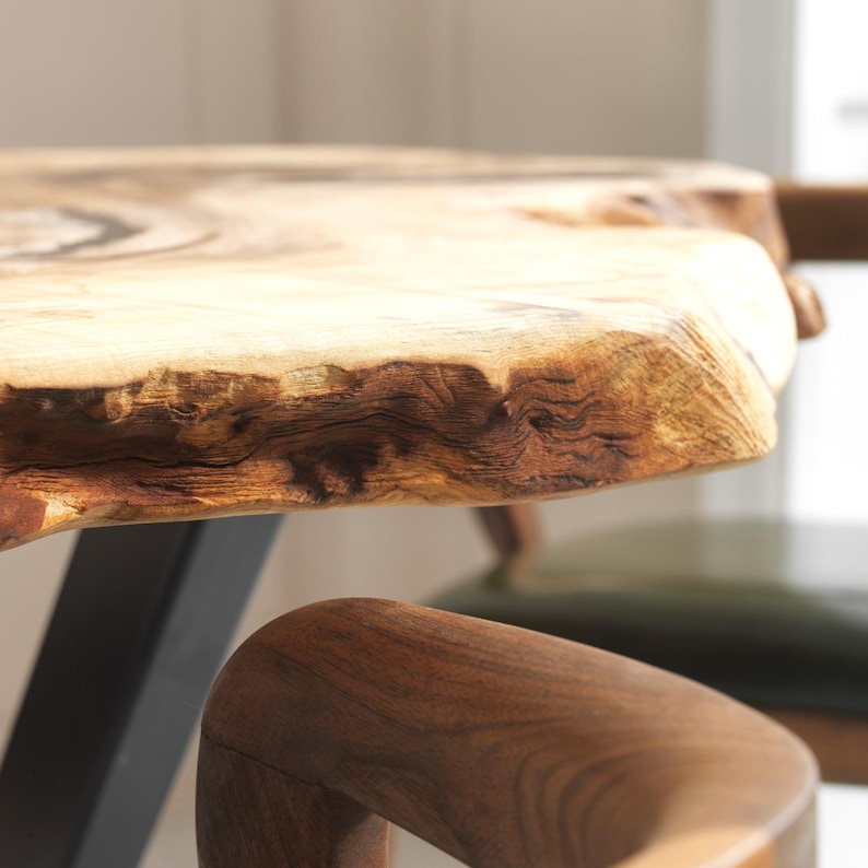 May include: A close-up of a wooden table with a natural edge. The table has a light brown surface with darker brown details. A wooden chair with a curved armrest is in the foreground. The table is supported by a black metal leg.