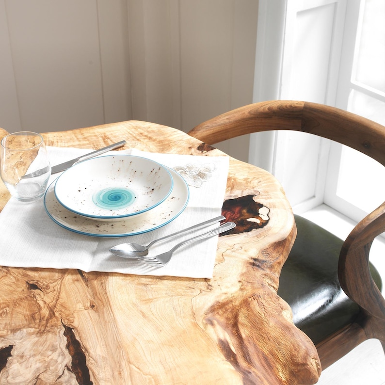 May include: A table setting featuring a white plate and bowl with a blue swirl design, a knife, fork, and spoon. The table is made of wood and has a natural, organic shape. A glass of water and a white napkin are also present.