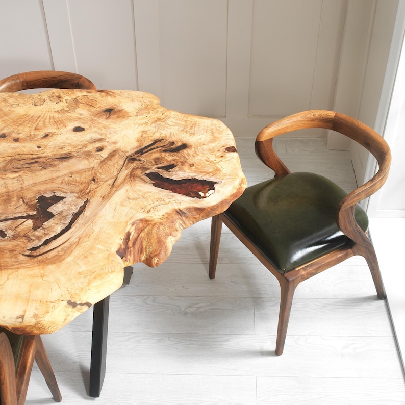 May include: A live edge wooden table with a natural, light-colored finish and dark metal legs. A wooden chair with a curved back and a green leather seat is next to the table. The table's dimensions are approximately 48 inches long and 30 inches wide.