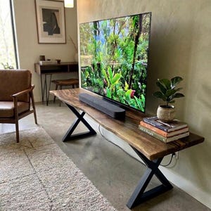 Può includere: Un mobile TV in legno con bordi naturali e gambe in metallo nero. Un televisore a schermo piatto mostra una scena della giungla. Una soundbar è sotto la TV. Una piccola pianta in vaso e libri sono sul mobile.