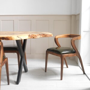May include: A wooden dining table with a live edge and black metal legs is paired with wooden chairs featuring dark green leather seats. The chairs have curved backs and slender legs, creating a modern, natural aesthetic.