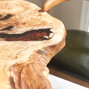May include: Close-up of a live edge wooden table with a natural, organic shape. The wood grain is visible, with a dark red and black resin-filled void. A green leather chair is partially visible in the background.