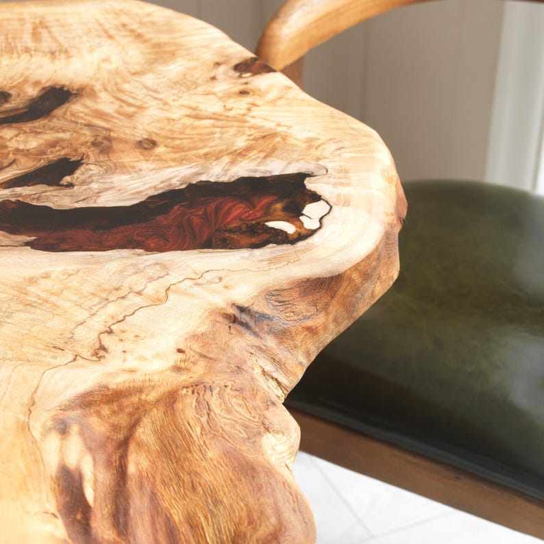 May include: Close-up of a live edge wooden table with a natural, organic shape. The wood grain is visible, with a dark red and black resin-filled void. A green leather chair is partially visible in the background.