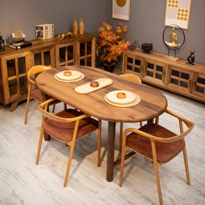 Puede incluir: Una mesa de comedor ovalada de madera con cuatro sillas. La mesa es de color marrón medio y tiene una sola pata central. Las sillas tienen marcos de madera y asientos tapizados de color óxido. La mesa está puesta con manteles individuales y cuencos pequeños de madera.