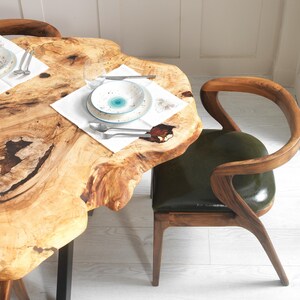 May include: A dining table with a natural wood slab top, paired with a wooden chair featuring a green leather seat. The table is set with white plates, silverware, and a clear glass. The table's edge has a unique, organic shape.