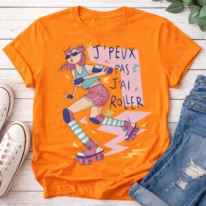 Op de afbeelding: Oranje t-shirt met een cartoon rolschaatser ontwerp, roze haar en zonnebril. De Franse tekst "J'peux pas, j'ai roller" is afgebeeld. Het shirt wordt gecombineerd met een blauwe jeans en witte sneakers.