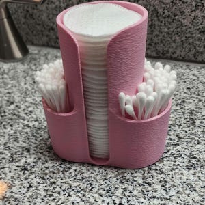 May include: A pink, 3D-printed organizer for cotton pads and swabs. The organizer has a tall, central compartment for cotton pads and two side compartments for cotton swabs. The organizer is sitting on a speckled countertop.