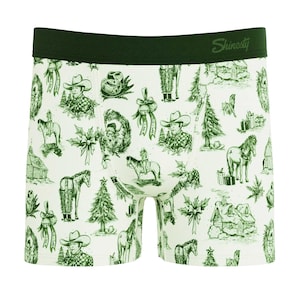 Puede incluir: Calzoncillos bóxer blancos con cintura verde y un estampado repetido de ilustraciones de vaqueros, caballos, árboles de Navidad y cajas de regalo. El texto "Shinesty" está impreso en la cintura.