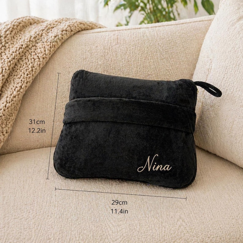 Puede incluir: Una almohada de felpa negra, de 31 cm de alto y 29 cm de ancho, con el nombre "Nina" bordado en escritura dorada. La almohada tiene una correa y un lazo. Est&aacute; sobre un sof&aacute; beige.