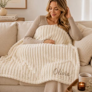Op de afbeelding: Een crèmekleurige, pluche plaid met een geribbelde textuur, voorzien van de naam Olivia geborduurd in een cursief schrift in de rechterbenedenhoek. De deken heeft een dikke, zachte en pluizige uitstraling met verticale ribbels.