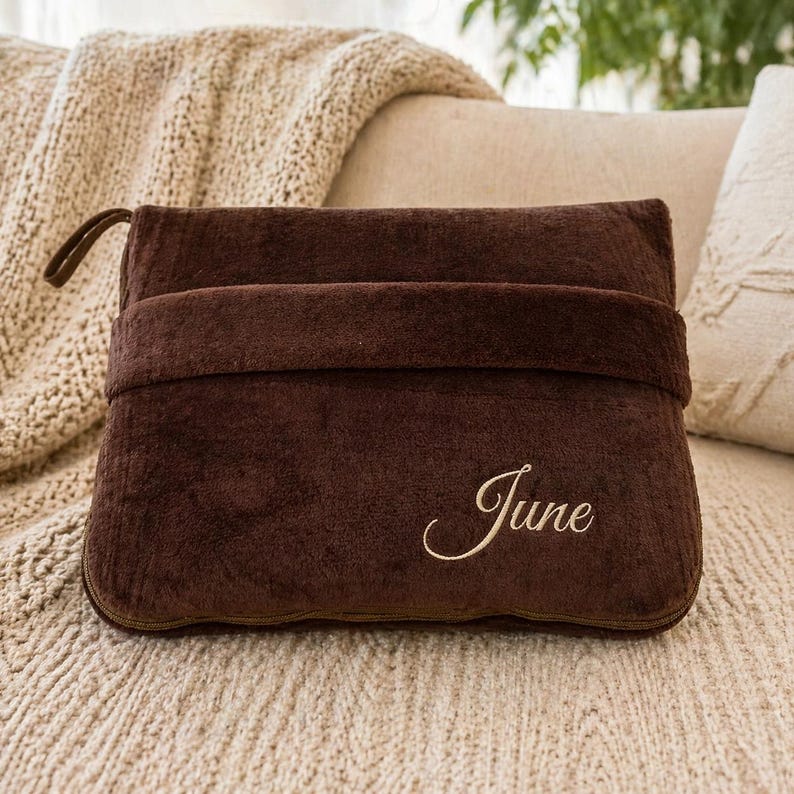 Puede incluir: Una almohada de felpa marr&oacute;n oscuro con una correa y un asa. La palabra "June" est&aacute; bordada en cursiva en la parte delantera. La almohada est&aacute; sobre una superficie beige texturizada.