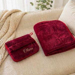 Puede incluir: Una manta burdeos doblada y un estuche de viaje a juego con el nombre "Olivia" bordado en escritura dorada. La manta es suave y afelpada, y el estuche tiene un asa de bucle. El conjunto se exhibe en un sof&aacute; color crema.