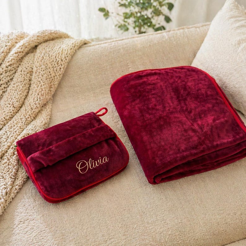 Puede incluir: Una manta burdeos doblada y un estuche de viaje a juego con el nombre "Olivia" bordado en escritura dorada. La manta es suave y afelpada, y el estuche tiene un asa de bucle. El conjunto se exhibe en un sof&aacute; color crema.