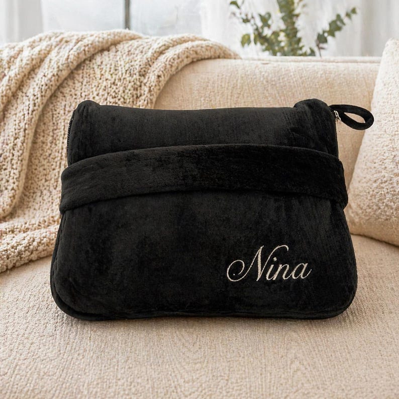 Puede incluir: Una almohada de viaje negra de felpa, con una correa envolvente y cierre de cremallera. El nombre "Nina" est&aacute; bordado en cursiva en la parte delantera. La almohada est&aacute; sobre un sof&aacute; beige.